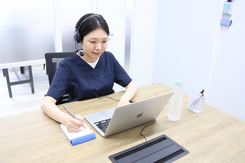 デスクでノートパソコンを開き、画面に向かって笑顔で話しかけているドクターメイトのオンコールナースの女性の写真。インカムをつけ、左手でパソコンを操作しながら、右手ではペンを持ってメモを取っています